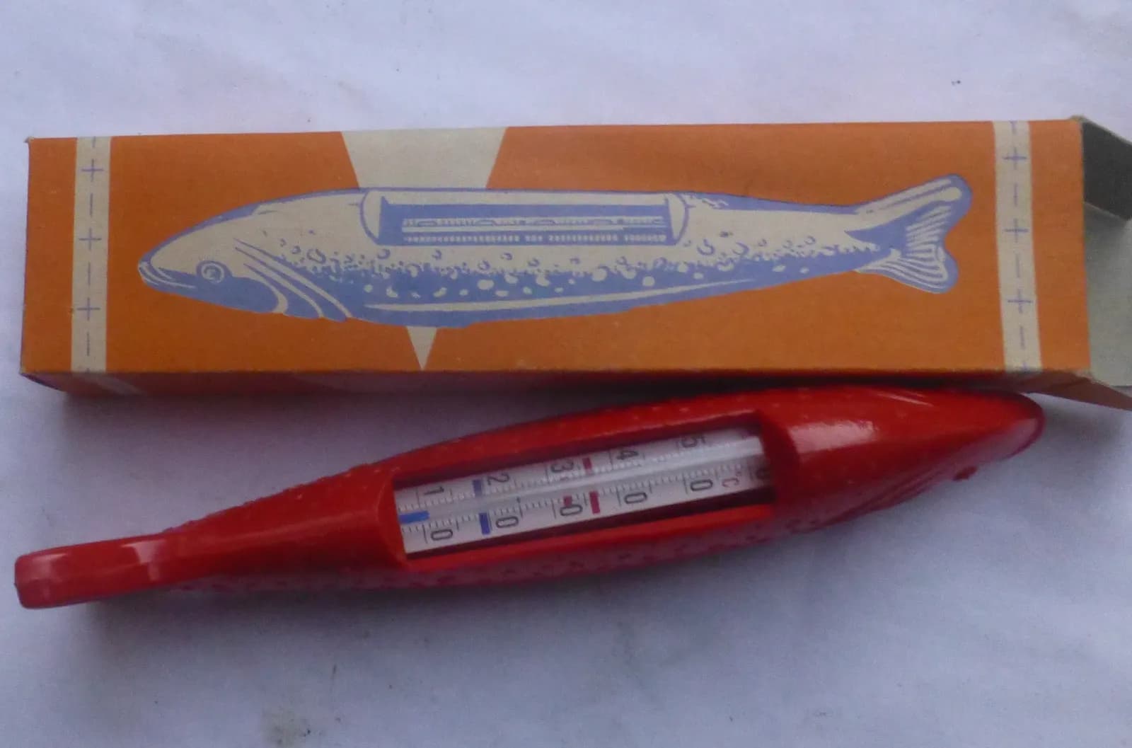 Thermomètre de bain rouge en forme de poisson, avec sa graduation en degrés, posé devant sa boîte orange illustrée d'un poisson