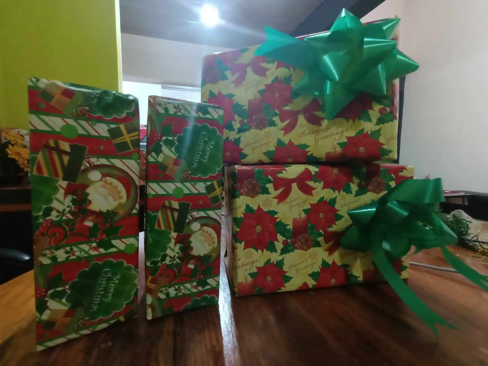 Quatre paquets cadeaux emballés de papier de Noël rouge, vert et jaune, avec de gros nœuds verts, posés sur une table en bois