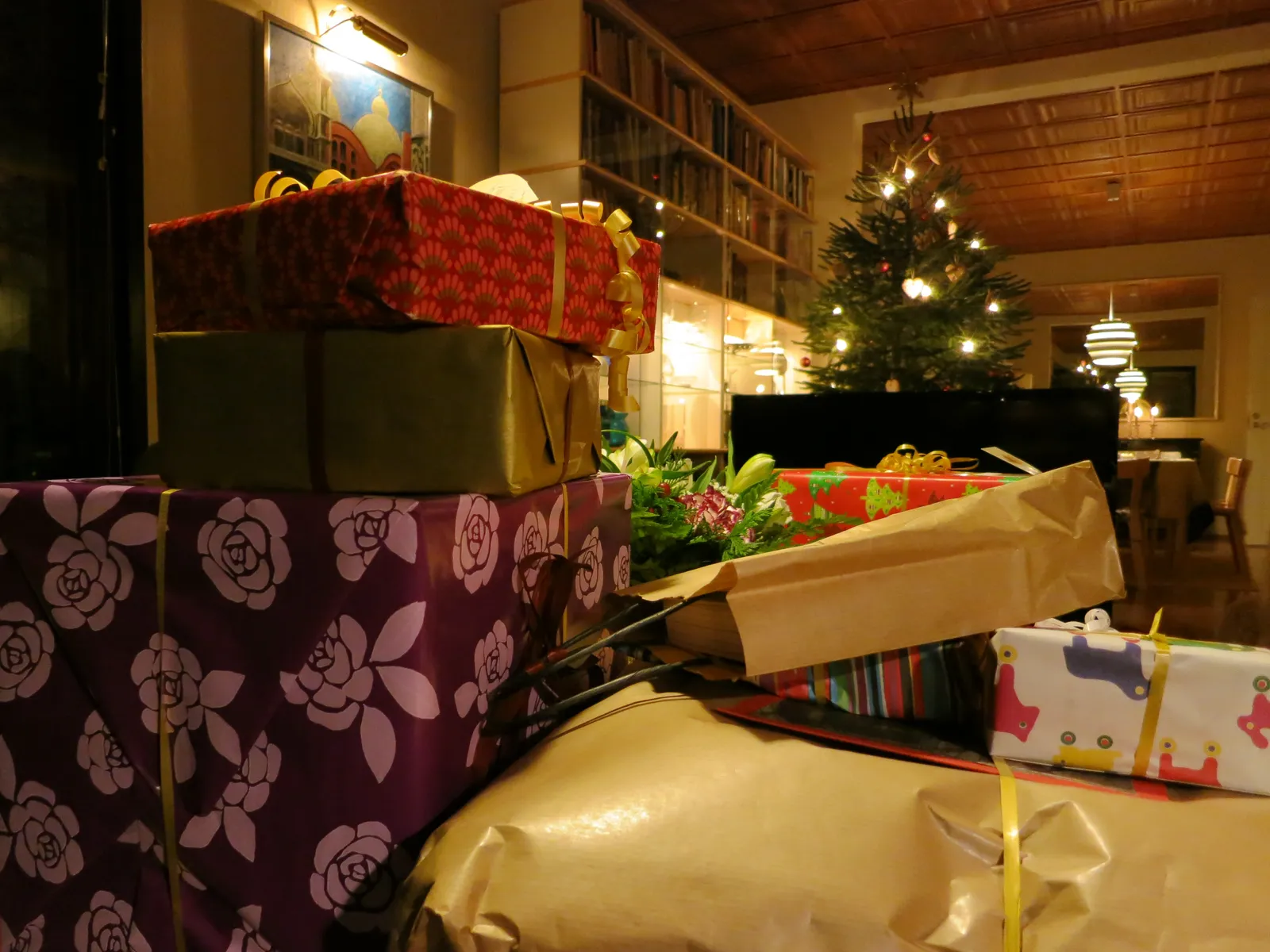Paquets cadeaux emballés de papiers colorés empilés dans un salon, avec un sapin de Noël illuminé au fond