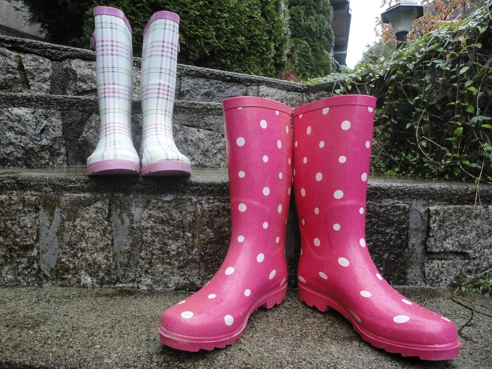 Deux paires de bottes de pluie, l'une rose à pois blancs, l'autre à carreaux, posées sur des marches en pierre mouillées