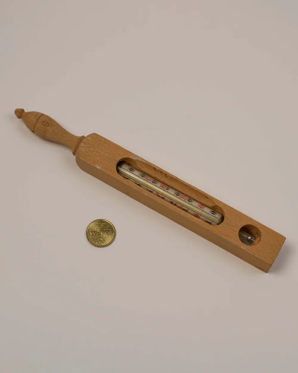 Ancien thermomètre de bain en bois clair, avec sa graduation sous verre et un manche tourné, posé à côté d'une pièce de monnaie
