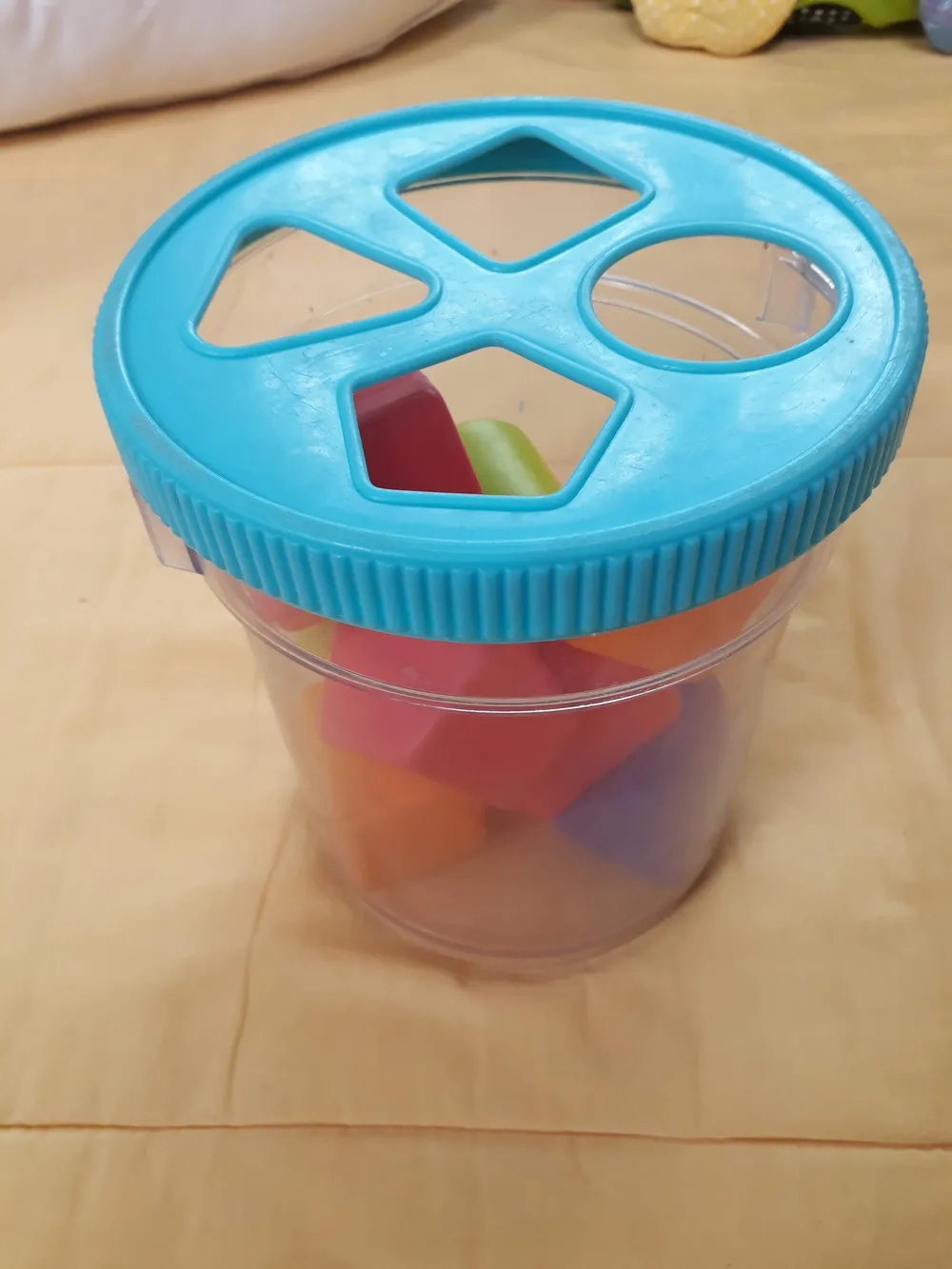 Boîte à formes en plastique transparent avec un couvercle bleu percé d'un triangle, d'un carré, d'un rond et d'un pentagone, remplie de blocs colorés